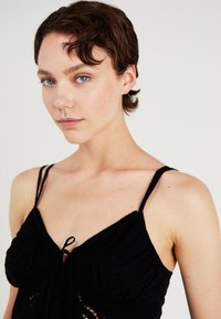 Svart camisole med tynne stropper, broderte detaljer, knyttet frontlukking og teksturert stoff. Nærbilde som fokuserer på utringningen.