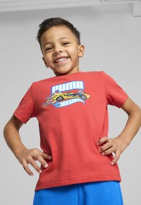 Camiseta de algodón roja con un gráfico de un coche amarillo con llamas, acompañado del texto "PUMA" y "Hot Wheels". Manga corta, cuello redondo.
