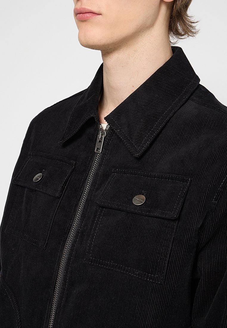 Veste en velours côtelé noir avec un col, fermeture éclair à l'avant et deux poches poitrine avec boutons-pression. Le tissu texturé est visible.