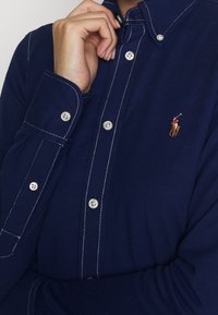 Person wearing navy blue buttoned shirt with visible white stitching and a small embroidered polo player logo on the chest.