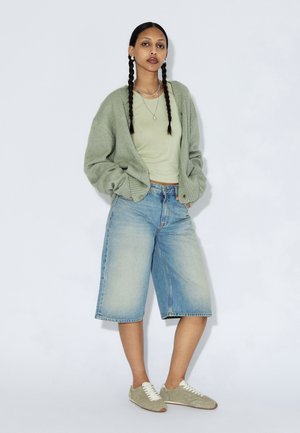 Cárdigan verde claro sobre una camiseta verde claro, combinado con pantalones culottes de mezclilla azul y zapatillas deportivas grises. Peinado con trenzas y collares en capas.