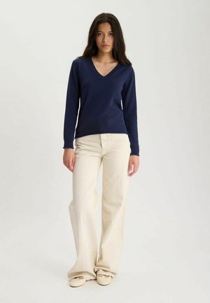 Femme debout portant un pull bleu marine à col en V, un pantalon large crème et des chaussures claires sur un fond uni.
