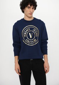 Tamsiai mėlynas džemperis su apvalia iškirpte, pagamintas iš medvilnės, su auksiniu apvaliu logotipu: "VERSACE JEANS COUTURE" ir centriniu "V". Lygus audinys, laisvas fasonas.