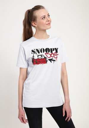 Ung kvinde med hestehale iført en hvid Snoopy hund T-shirt og sorte bukser, kigger op med et let smil mod en ensfarvet baggrund.