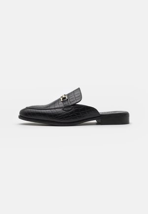 Mocasín negro de cuero tipo slip-on con textura de cocodrilo y detalle de hebilla metálica dorada, tacón bajo y punta estrecha.