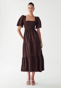 Robe maxi en beige à volants avec des manches courtes bouffantes, un corsage froncé et un ourlet à volants. Portée avec des sandales blanches à lanières. Texture de tissu lisse.