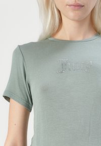 Īsām piedurknēm, gaiši zaļš T-krekls mīkstā audumā, kurā vidū ir akmeni rotāta "Juicy" logo. Šuves detaļas uz piedurknēm.