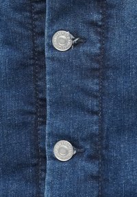 Tumed sinine denimpind, millel on silmapaistvad õmblused. Kahe hõbedase nupuga, millel on graveeritud tekst, on nähtavad, lisades riietusele detaili.