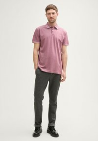 Rød polo t-shirt med hvidstribede kraver, sammen med mørkegrå teksturerede bukser og sorte støvler. Modellen står på en neutral baggrund.