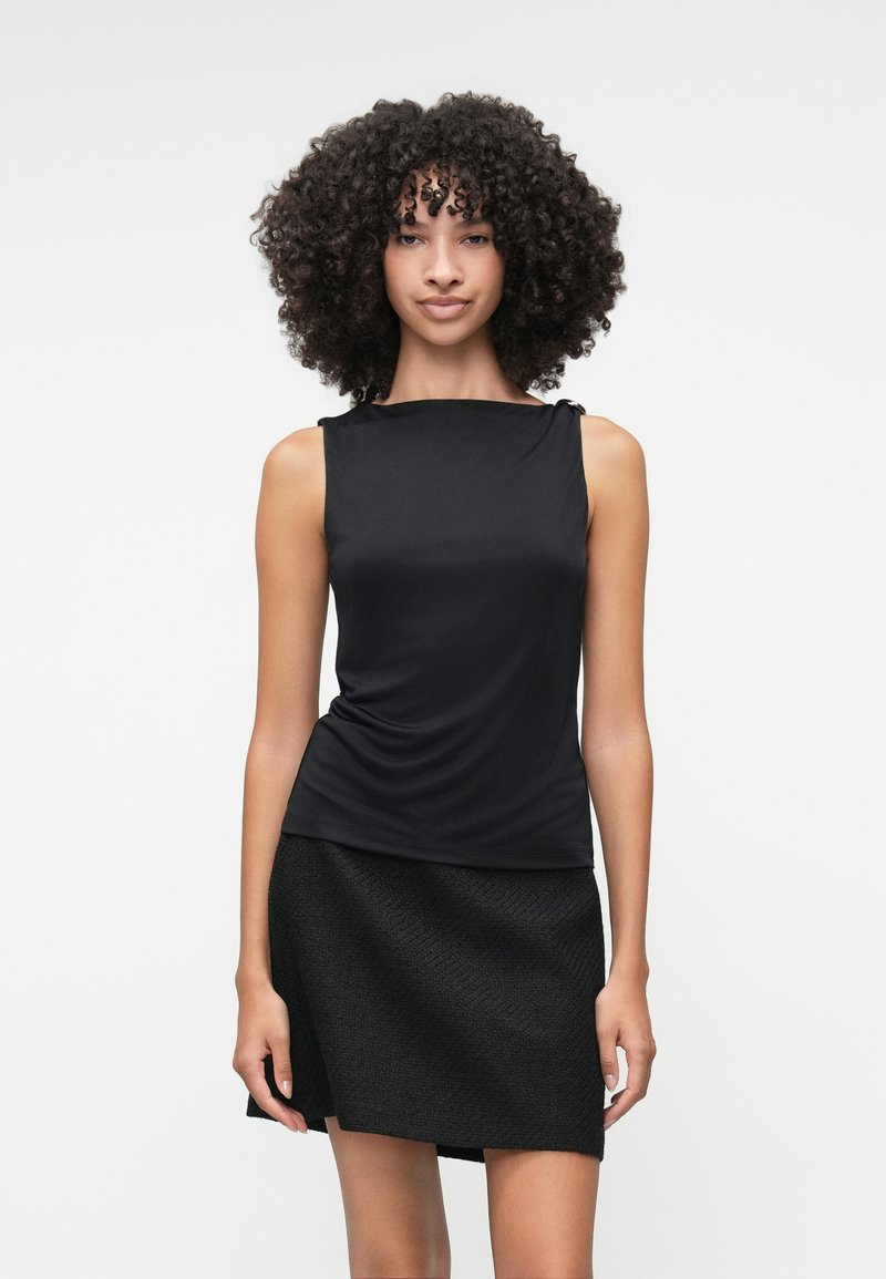 Μαύρη, αμάνικη μπλούζα με ελαφριά ρίσκα και γωνιακό ντεκολτέ, σε συνδυασμό με μια υφασμάτινη μαύρη ασύμμετρη φούστα. Απλός σχεδιασμός, χωρίς ορατές διακοσμήσεις.