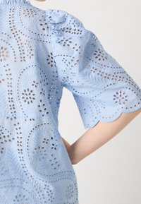 Gros plan sur un chemisier en tissu œillet bleu clair avec broderie découpée de fleurs et de feuilles, manches bouffantes mi-longues au niveau du coude.