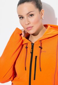 Oranžová kapucínová bunda vyrobená z měkkého materiálu, s černým zipem, šňůrkami a vytištěným nápisem "sport" v purpurové barvě.