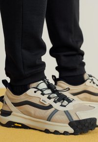 Beige och svart sneakers med texturerat tyg, tonala överlägg, svarta snören och Vibram-sula, tillsammans med svarta joggers.