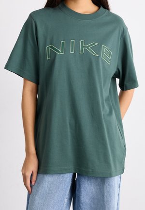 Personne portant un t-shirt Nike ample vert foncé à manches courtes et un jean en denim bleu clair, debout devant un fond uni.