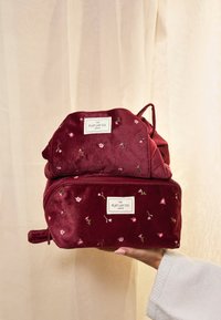 Mochila de terciopelo burdeos con patrón floral bordado, dos compartimentos y dos correas de tela, que presenta la etiqueta "The Flat Lay Co."