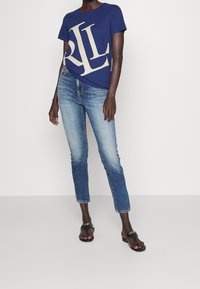 Žena v modrém tričku s velkým potiskem, světle modrých slim-fit džínách a černých plochých sandálech se zlatými detaily na bílém pozadí.