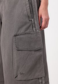 Pilkos krovinių kelnės, pagamintos iš tvirto audinio, su šoniniu kišenėle, trimis juostelėmis šonuose ir mažu prekės ženklo logotipu, įspaustu ant kišenės.