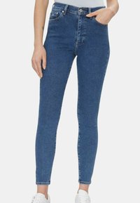 hoog-middelhoge, skinny jeans in donkerblauwe denim met stiksel-details, vijf zakken en een knoopsluiting, gecombineerd met witte sneakers.