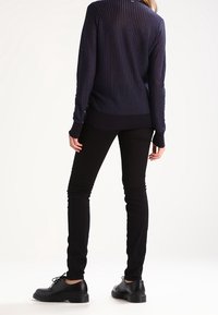 Marinblå, ribbad tröja med hög halsringning, kombinerad med smala svarta byxor och svarta snörskor, sedd bakifrån.