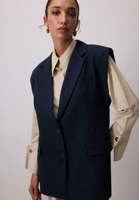 Námornícky modrý bezrukávový blejzer s dvoma gombíkmi a bočnými vreckami, nosený cez svetlobežový košeľový tričko s golierom. Hladká tkanina so štruktúrovaným dizajnom.