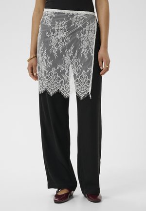 Tablier en dentelle florale blanche porté sur un pantalon noir à jambes larges avec des chaussures plates bordeaux, visible de la taille aux pieds.