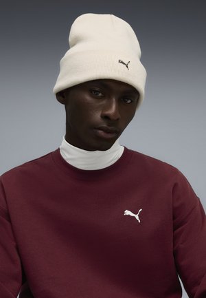 Hvid strikket hue, maroon sweatshirt med rund hals, prydet med et hvidt Puma-logo på brystet, sat mod en gradient grå baggrund.