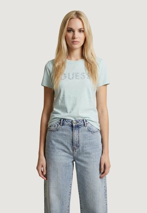 Mladá žena s dlouhými blond vlasy, která má na sobě světle modré tričko Guess a vysoké světle modré džíny, stojí před jednobarevným pozadím.