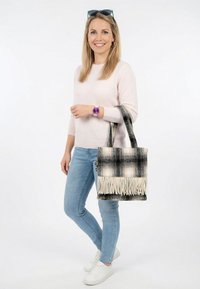 Mujer sonriente con suéter rosa, jeans azul claro y zapatillas blancas, llevando un bolso tote con flecos a cuadros blancos y negros en el brazo derecho.