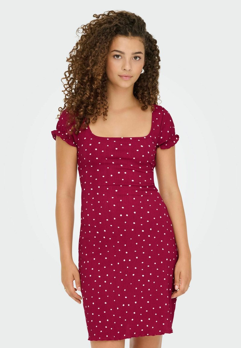 Jeune femme aux cheveux frisés portant une robe courte à manches courtes de couleur bordeaux avec des pois blancs, se tenant devant un fond uni.