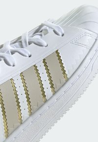Bílé tenisky s klikatým zlatým a krémovým vzorem, texturovanou gumovou podrážkou a bílými tkaničkami. Hladký kožený materiál.