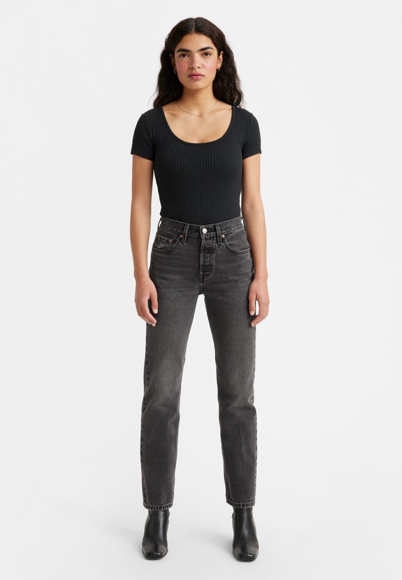 Bodysuit negro con trenzado y escote redondo, combinado con jeans de mezclilla gris oscuro de tiro alto. La modelo lleva botas de tobillo y está de pie contra un fondo blanco.