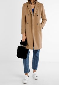 Jasnobrązowy płaszcz z mieszanki wełny z dwurzędowym zapięciem na czarne guziki, w połączeniu z niebieską bluzką, dżinsami, czarną torebką z futerkiem oraz białymi butami.