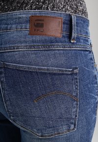 Jeans de denim azul con un parche de cuero en la cintura, que presentan bolsillos traseros con costuras curvas y un acabado desgastado.
