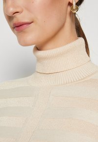 Beige Rollkragenpullover mit geripptem Design und strukturierten Streifen. Glatter Stoff mit leicht glänzender Oberfläche und taillierter Silhouette.