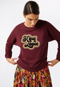 Bordeaux sweatshirt met lange mouwen, raglan ontwerp en het "King Louie" logo in zwart en goud. Het model draagt het in combinatie met een kleurrijke geplooide rok.