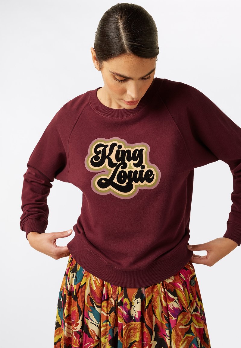 Bordeaux sweatshirt met lange mouwen, raglan ontwerp en het "King Louie" logo in zwart en goud. Het model draagt het in combinatie met een kleurrijke geplooide rok.