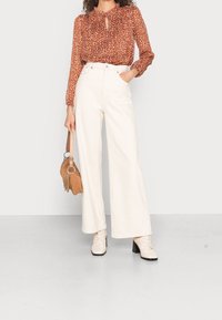 Blouse à manches longues de couleur rouille avec motif floral, détail en forme de clef à l'encolure ; pantalon large crème ; sac à main beige ; bottines beige.