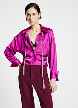 Blouse en satin fuchsia avec col cranté, coupe oversize et fermeture à boutons sur le devant. Comprend un détail décoratif avec un pompon argenté.