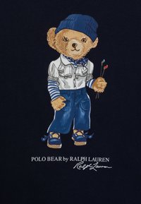 Ilustrace polo medvěda na námořnickém pozadí, oblečeného v bílém tričku s volány, modrých džínách a modrých botách, držícího štětce.