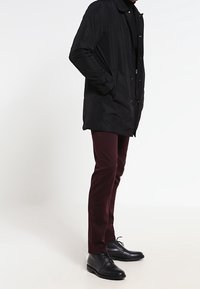 Černá lehká bunda s límcem a zapínáním na knoflíky, nošená přes černou košili. Kombinováno s vínovými kalhotami a černými botami.