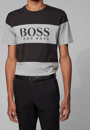 Man draagt een zwart-grijs Hugo Boss T-shirt met de tekst "BOSS HUGO BOSS", zwarte broek en zwarte riem tegen een grijze achtergrond.