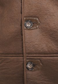 Nahaufnahme von braunem Wildlederstoff mit zwei großen runden Knöpfen, die an Lederlaschen entlang einer vertikalen Naht angenäht sind.