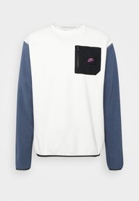 Vit sweatshirt med marinblå ärmar, med en svart dragkedjeficka på bröstet och en lila Nike-logotyp. Slät tyg, design med rund halsringning.