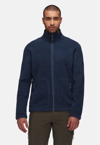 Navy fleece jacket with a high collar, full front zipper, and two side pockets. Features subtle logo detail on the chest and textured fabric.