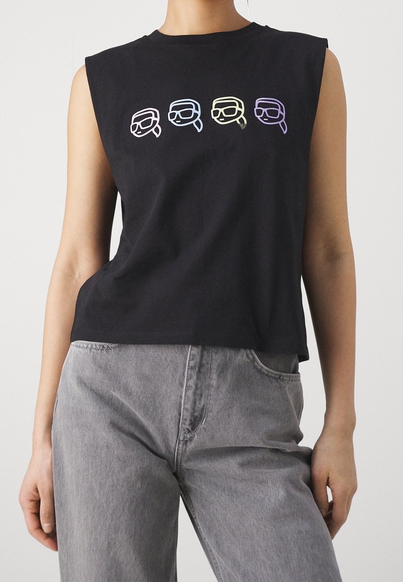 Chemise noire sans manches avec quatre petits visages colorés en contour portant des lunettes sur la poitrine, associée à un jean gris.