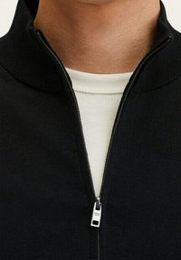 Sudadera negra con cremallera y cuello alto, con una cremallera plateada y una capa interior blanca. Tejido de punto texturizado.