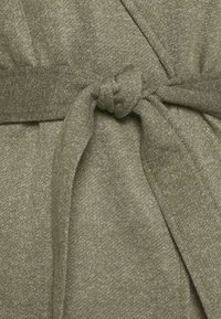 Olivově zelená pletená tkanina s texturovaným povrchem, která má zavázaný pásek. Tkanina má jemný lesk a měkký vzhled.