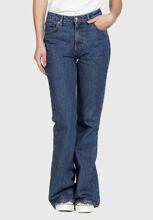 Donna che indossa jeans a vita alta, blu scuro, a zampa con cinque tasche e sneakers bianche, in piedi con una mano in tasca.