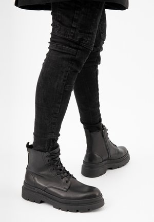 Stivali alla caviglia in pelle nera con punte rotonde, cuciture definite e suole in gomma spessa. Presentano un design con lacci e una zip laterale per una facile vestibilità.
