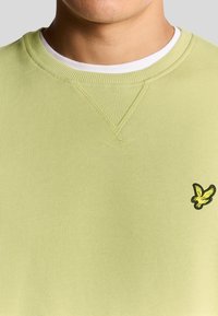 Felpa verde chiaro con texture a coste, scollatura rotonda e colletto interno bianco. Presenta un piccolo logo nero e giallo sul petto.